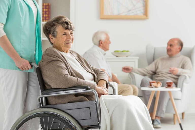 An older adult unpacks a box of personal belongings in a well-lit private room with supportive family members nearby.